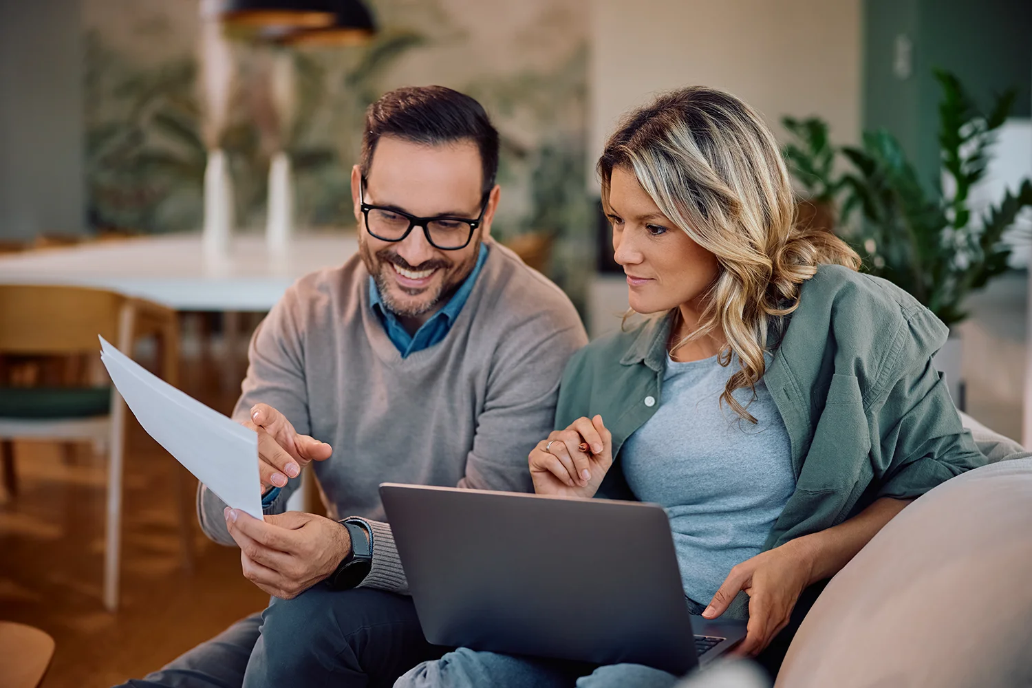 A man and woman sit together on a couch, looking at property tax documents and a laptop. The man is holding papers, smiling, and pointing at the documents, while the woman is sitting next to him, looking at the laptop. Both appear focused and engaged in a conversation about their property tax matters.